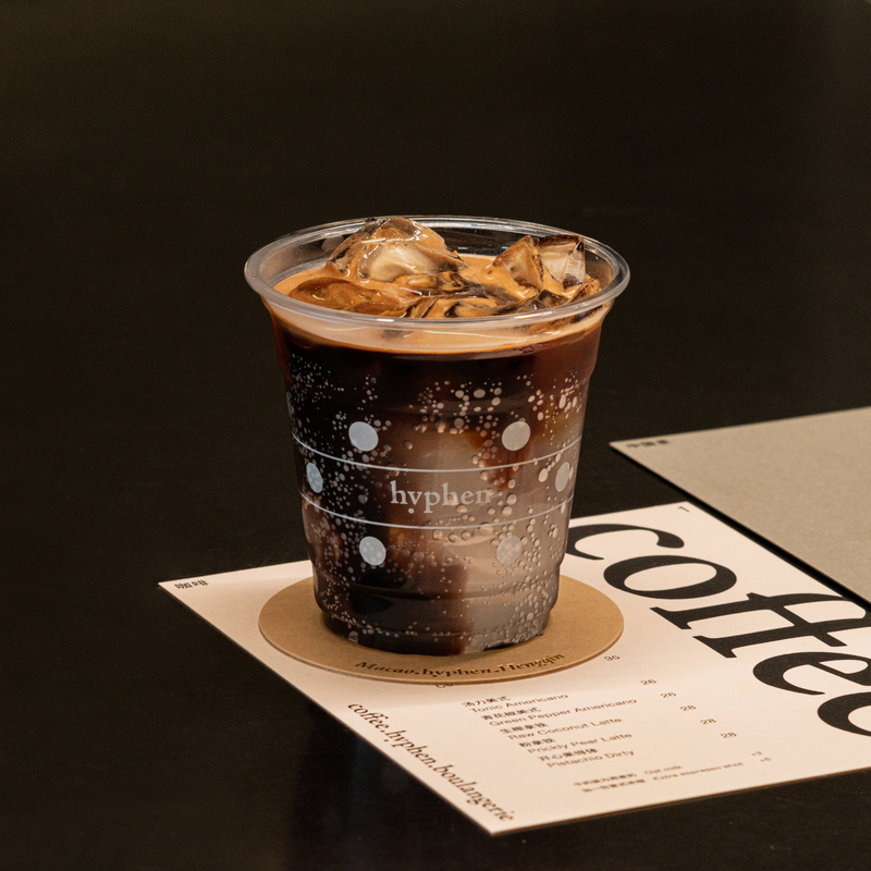 An iced americano in a branded hyphen. cup on a "Macau · hyphen · Hengqin" coaster, resting on the coffee menu at hyphen. Hengqin
