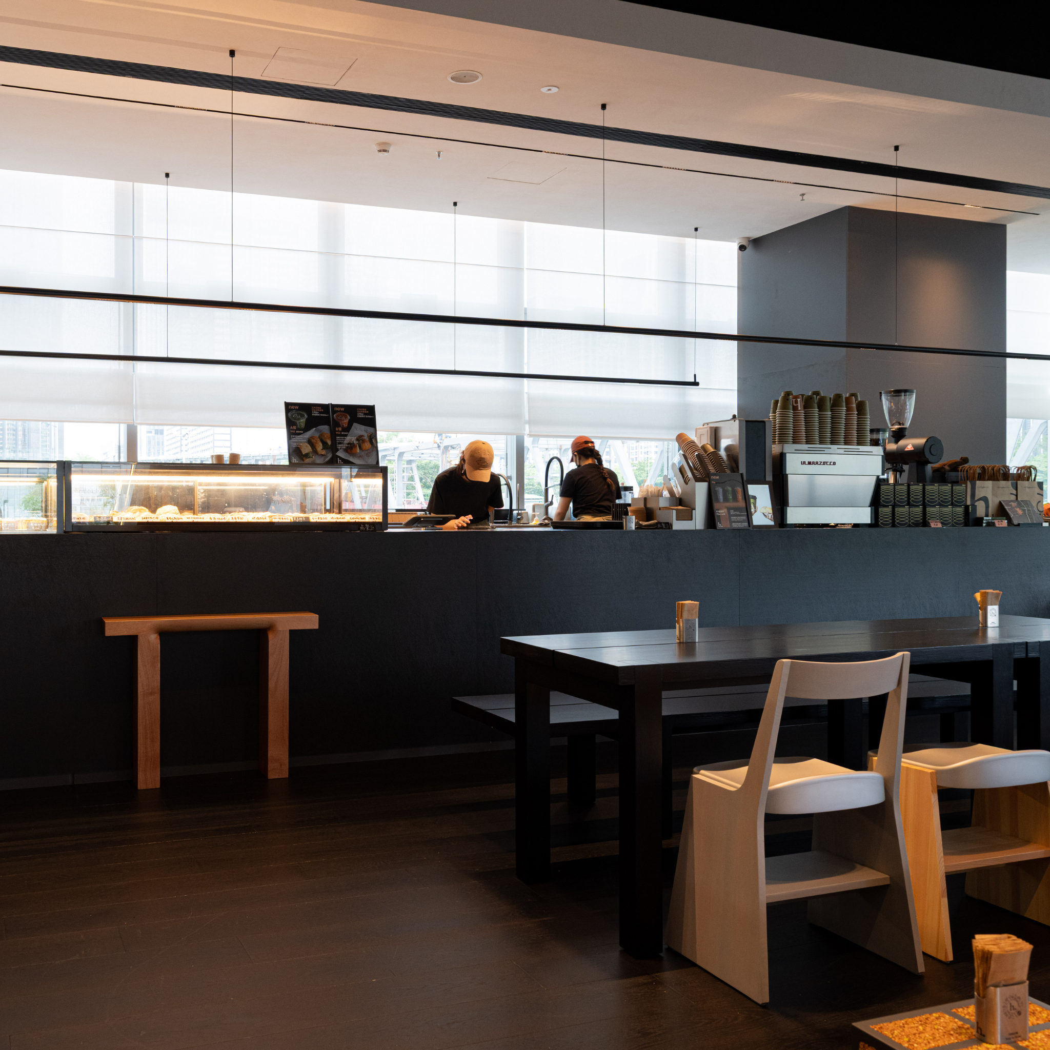 A modern cafe interior with a dark wood counter, two staff members behind the counter, and a dining area with dark tables and light wood chairs in the foreground.