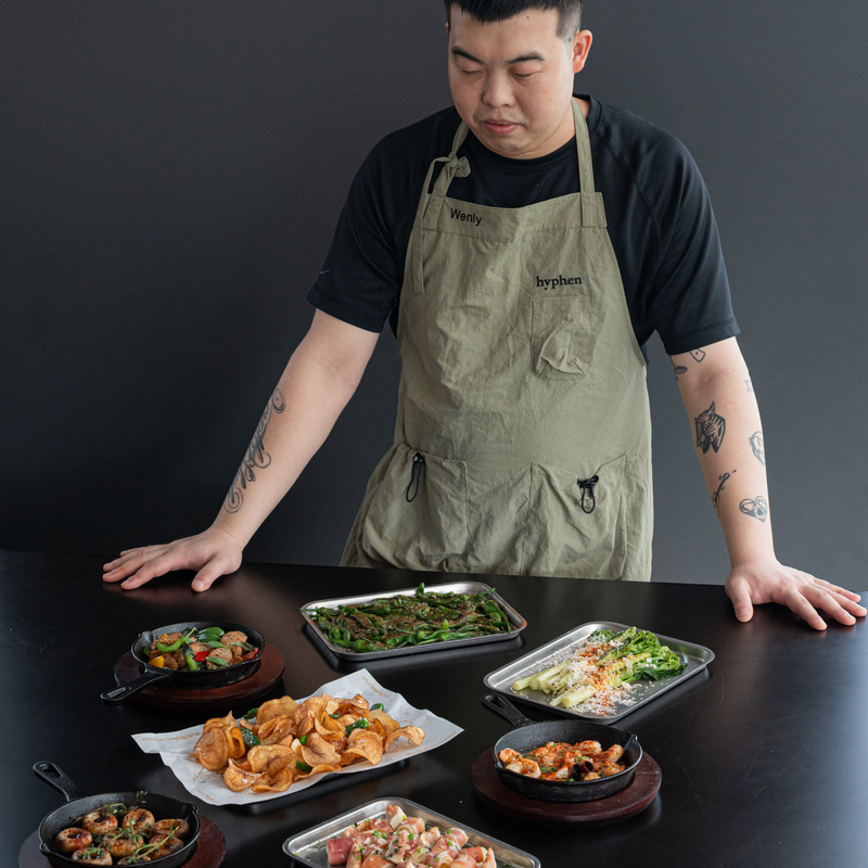 A man wearing a black t-shirt and an olive green apron stands behind a table displaying various prepared dishes in cast iron skillets and metal trays against a dark gray background.