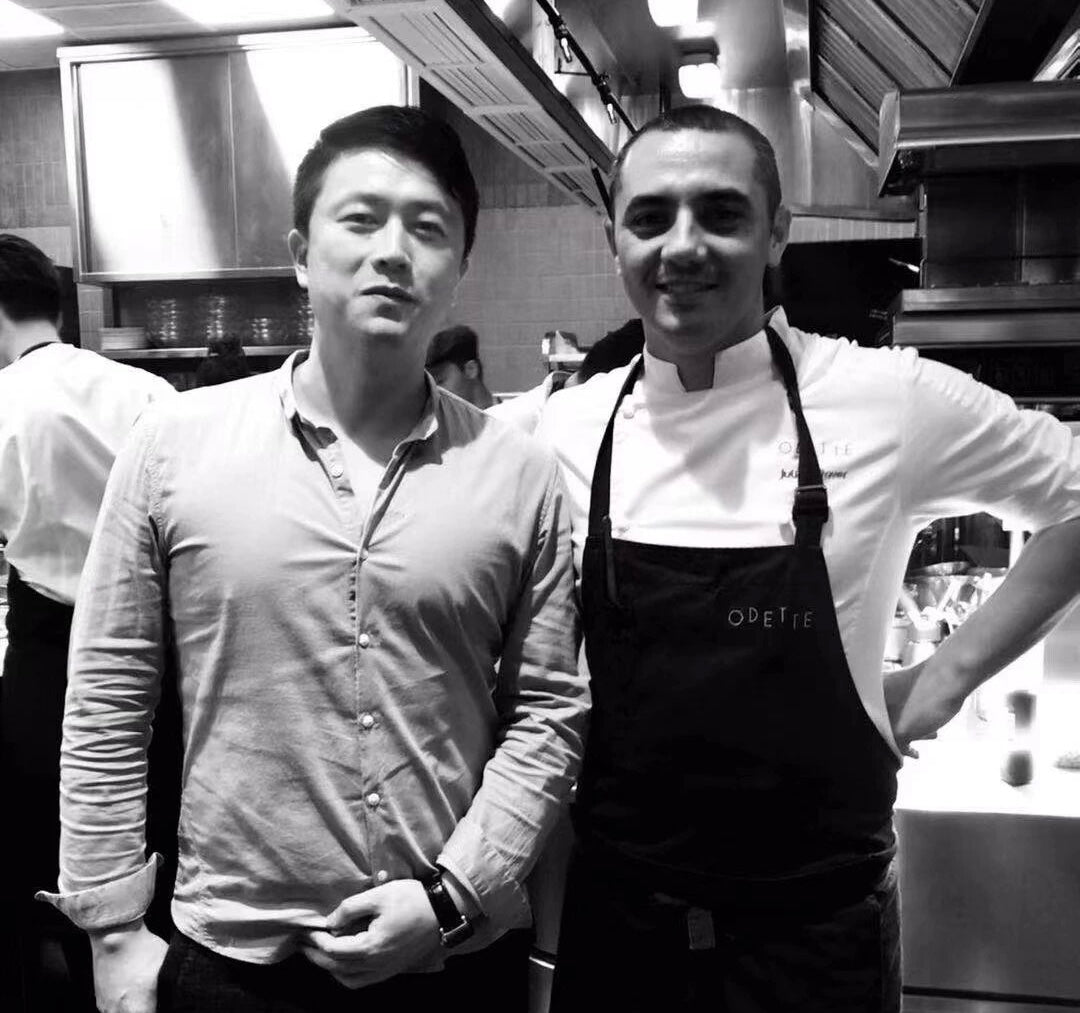 A black and white photo of two men standing together in a professional kitchen. The man on the right wears a white chef coat and a dark apron with the word 'OBETIE' visible on it, while the man on the left wears a light button-up shirt.
