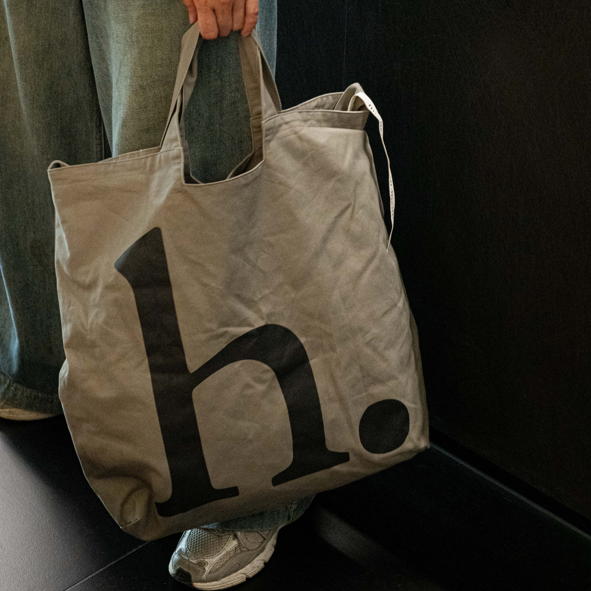 The hyphen. Everyday Tote in stone-grey canvas, carried by hand, with the oversized black "h." mark across the front