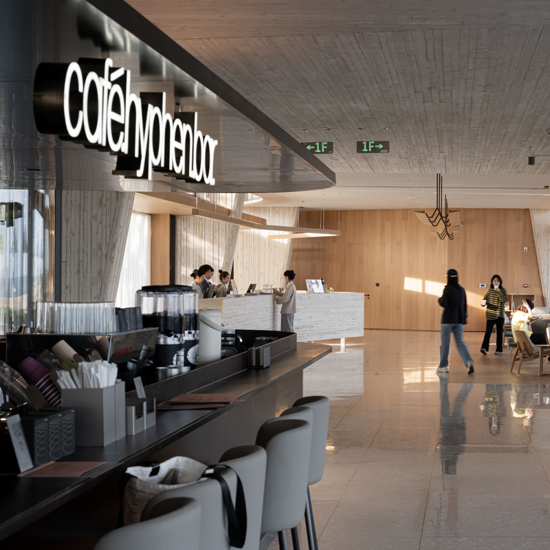 A modern cafe interior with a long bar counter, bar stools, espresso machines, and a large illuminated sign reading 'cafe hyphen bar'. Several people are visible in the space, and the room has light wood and concrete walls and ceiling.