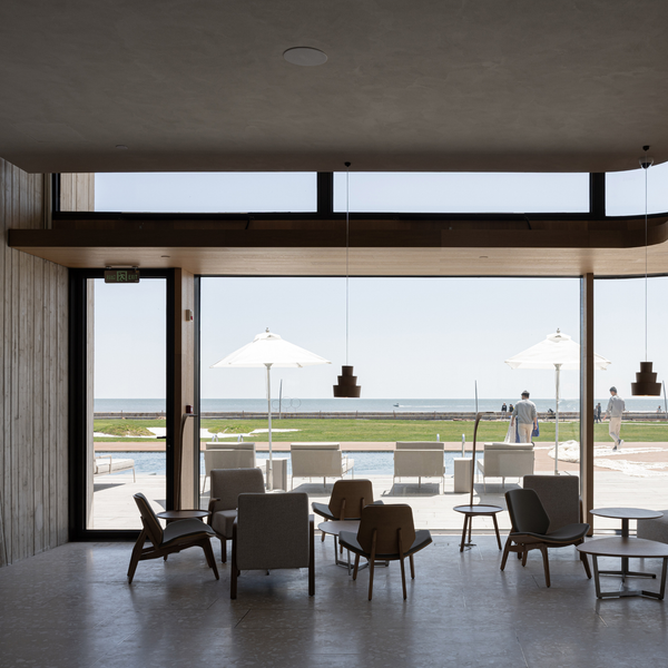 A modern open-plan interior with lounge chairs, tables, and pendant lights overlooking a pool and beachfront terrace. Large floor-to-ceiling windows and glass doors frame the outdoor view, with people visible in the distance near umbrellas and seating.