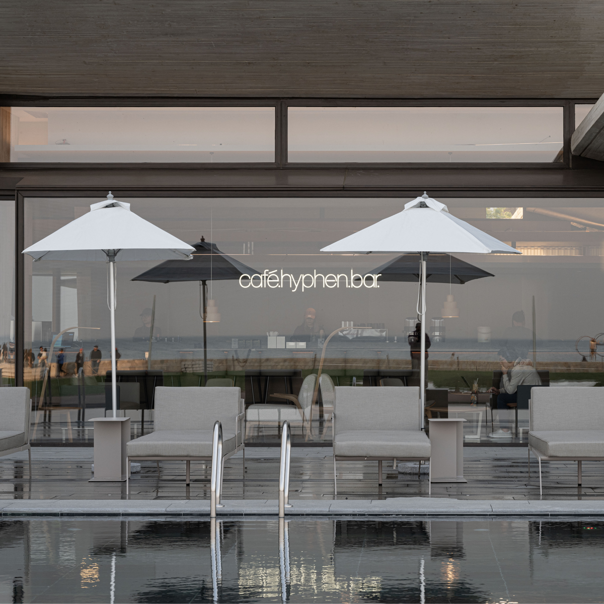 A modern outdoor seating area by a glass wall with two large white umbrellas, lounge chairs, and a visible 'cafehyphenbar' sign, with a pool in the foreground reflecting the scene.