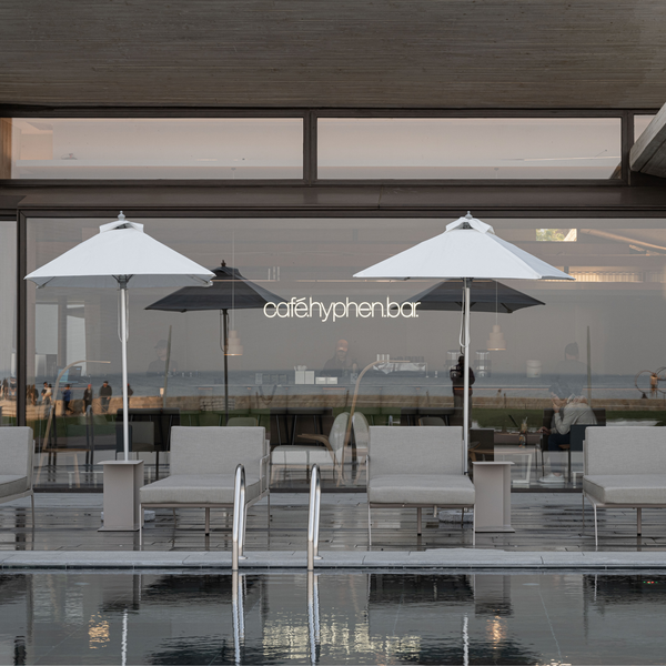A modern outdoor seating area by a glass wall with two large white umbrellas, lounge chairs, and a visible 'cafehyphenbar' sign, with a pool in the foreground reflecting the scene.