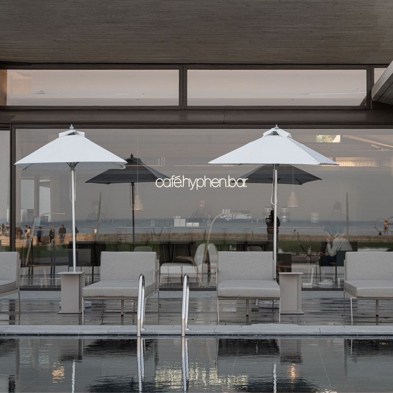 A modern outdoor seating area by a glass wall with two large white umbrellas, lounge chairs, and a visible 'cafehyphenbar' sign, with a pool in the foreground reflecting the scene.