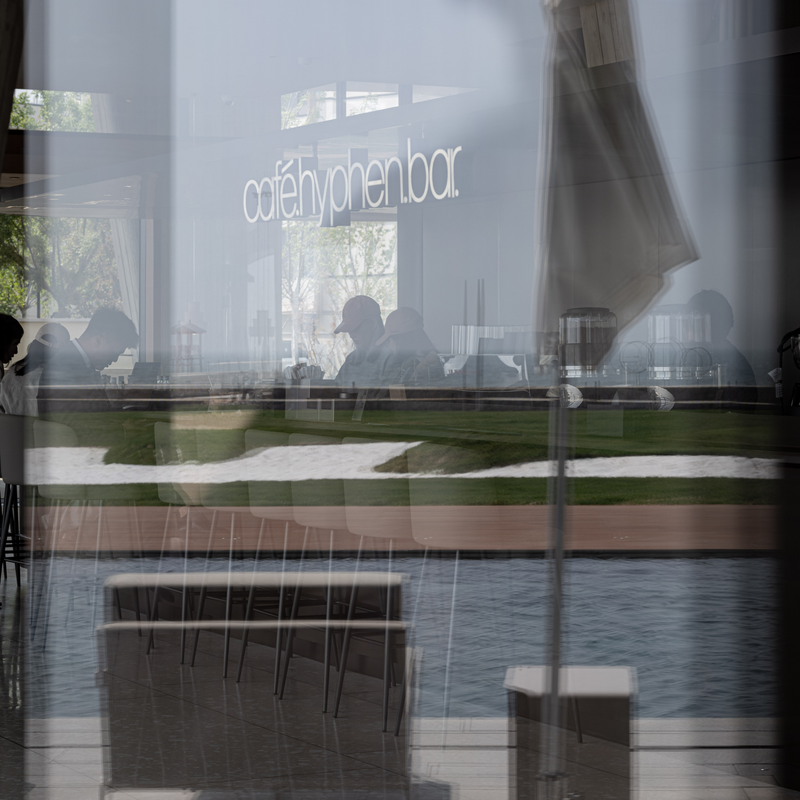 A reflective glass storefront of a modern cafe or bar, showing silhouettes of seated people, a white sign reading 'cafe hyphen bar', a green landscaped terrace, and water in the foreground.