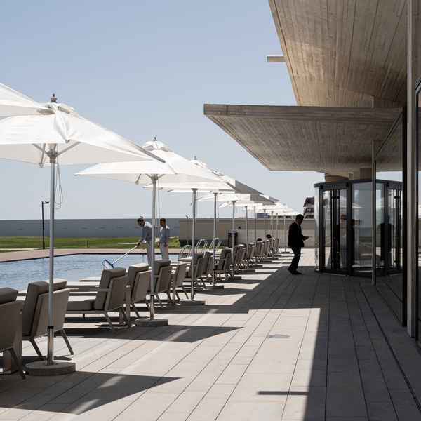 A modern outdoor poolside terrace with white patio umbrellas, lounge chairs, a light stone deck, and a large overhanging concrete roof. A person in dark clothing stands near the revolving glass doors, and two people are visible by the pool on the left.