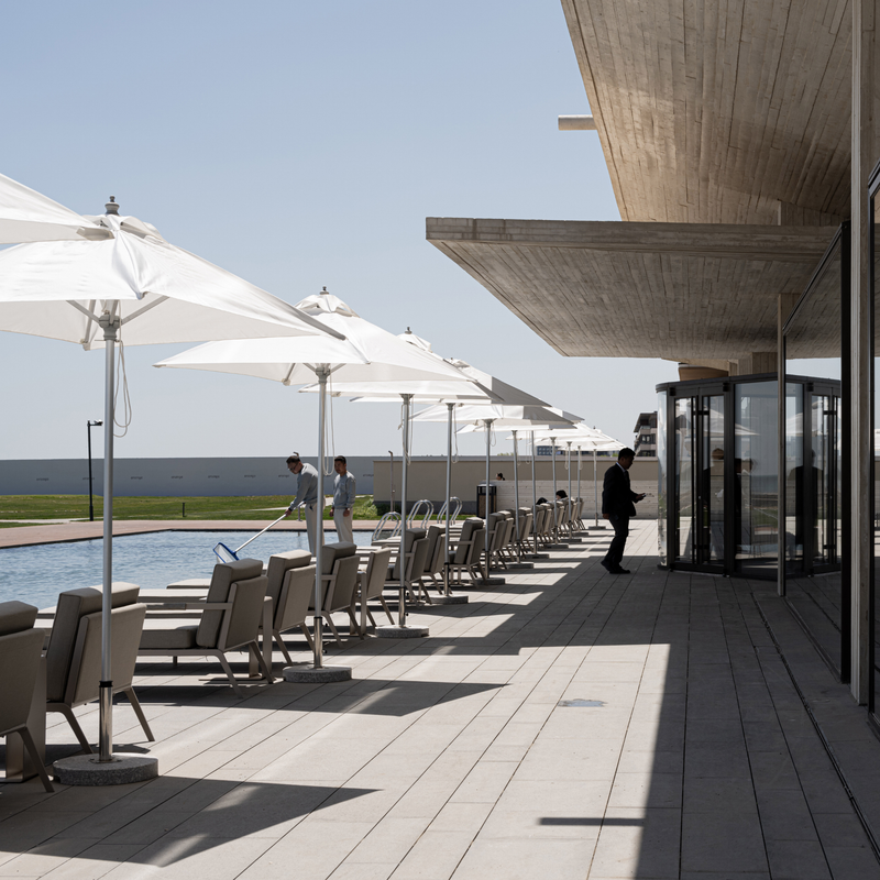 A modern outdoor poolside terrace with white patio umbrellas, lounge chairs, a light stone deck, and a large overhanging concrete roof. A person in dark clothing stands near the revolving glass doors, and two people are visible by the pool on the left.