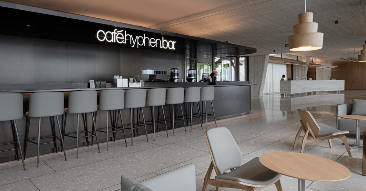 A modern cafe interior with a long bar counter, gray bar stools, hanging pendant lights, and light wood chairs with round tables. The wall displays the illuminated text 'cafe hyphen bar'.
