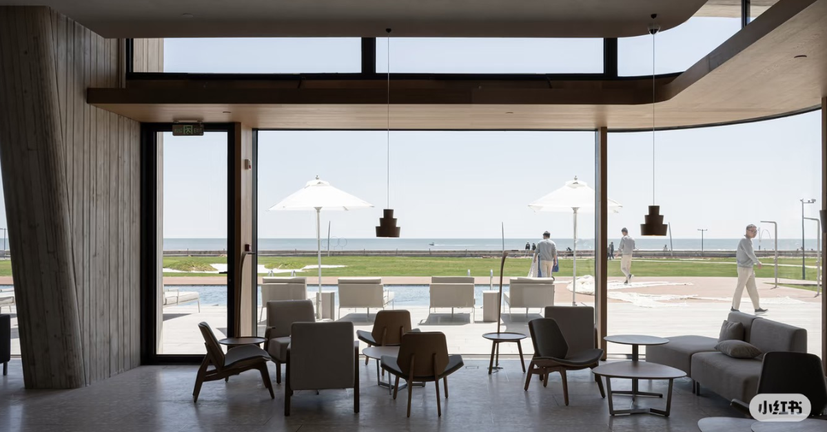 A modern interior lounge area with curved glass walls overlooking a beach and ocean, furnished with gray and brown chairs, tables, and a sofa. A few people are visible outside near umbrellas and poolside.