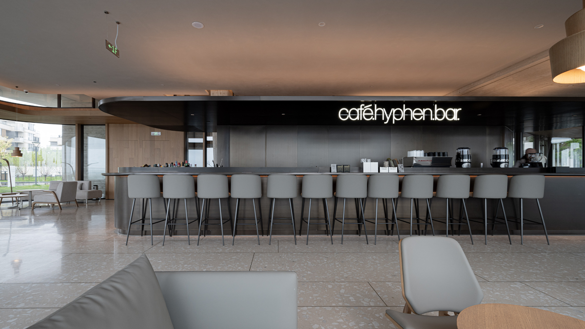 A modern interior bar scene with a long counter, multiple gray bar stools, and a black back wall featuring the white text 'cafehyphenbar'. The space has a warm neutral ceiling, tiled floor, and additional seating areas visible in the background.