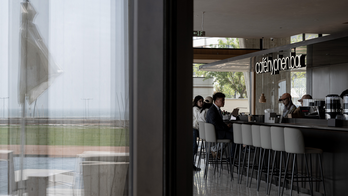 A view from inside a modern cafe looking out through a large window, with a bar counter, stools, and a few seated people visible in the background. The sign on the wall reads 'cafe phenobar'.