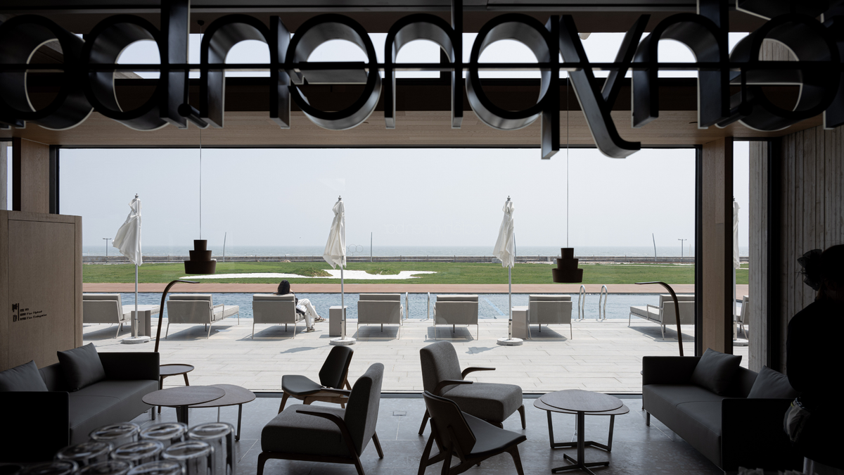 A modern interior lounge with large floor-to-ceiling windows overlooking a pool and outdoor seating area. The room features dark sculptural wall art, gray and black furniture, round tables, and a silhouetted figure near the right edge.