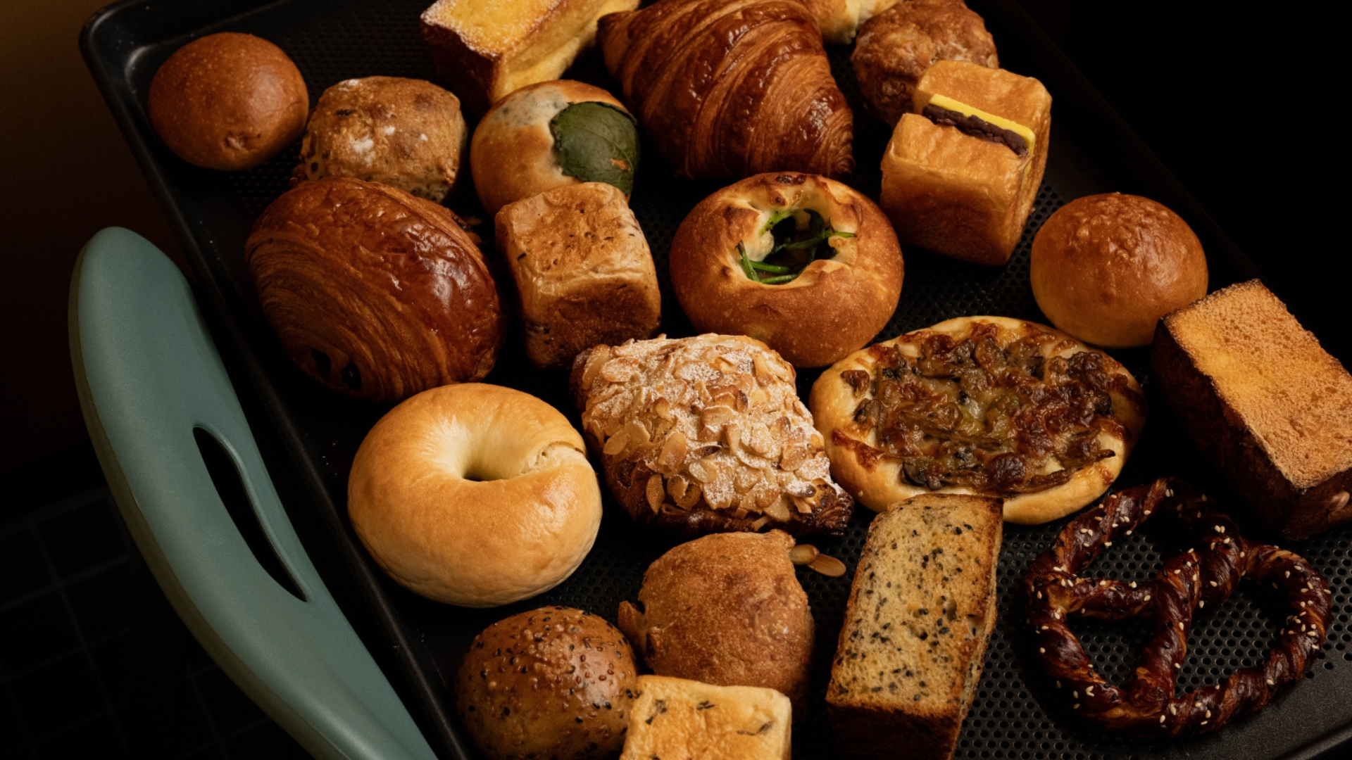 A variety of freshly baked breads, pastries, and sandwich fillings arranged on a black baking tray with a teal handle, photographed in warm, moody lighting.