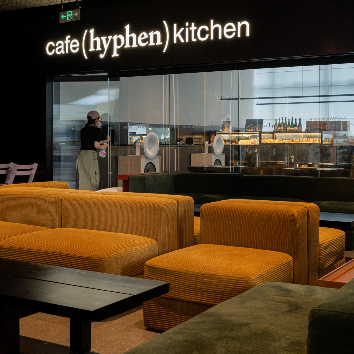 A modern cafe interior with mustard yellow corduroy upholstered seating in the foreground, a large glass window, and a visible person working behind it. The wall sign reads 'cafe (hyphen) kitchen'.