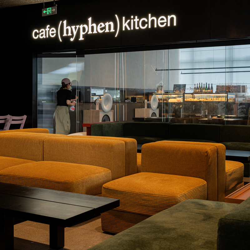 A modern cafe interior with mustard yellow corduroy upholstered seating in the foreground, a large glass window, and a visible person working behind it. The wall sign reads 'cafe (hyphen) kitchen'.