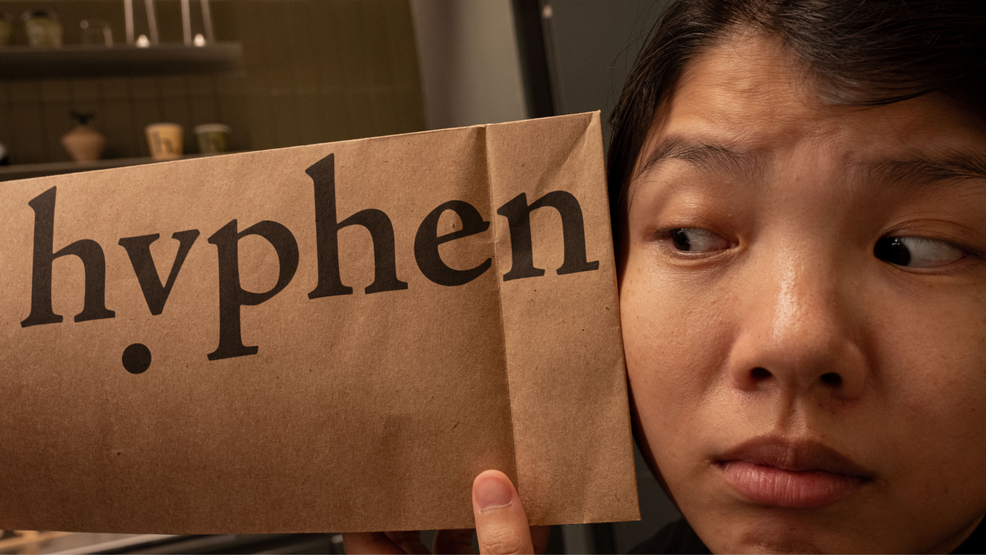 A close-up lifestyle shot of a person holding a brown paper bag with the brand name 'hyphen' printed in black serif font, with the person's face partially visible behind the bag.