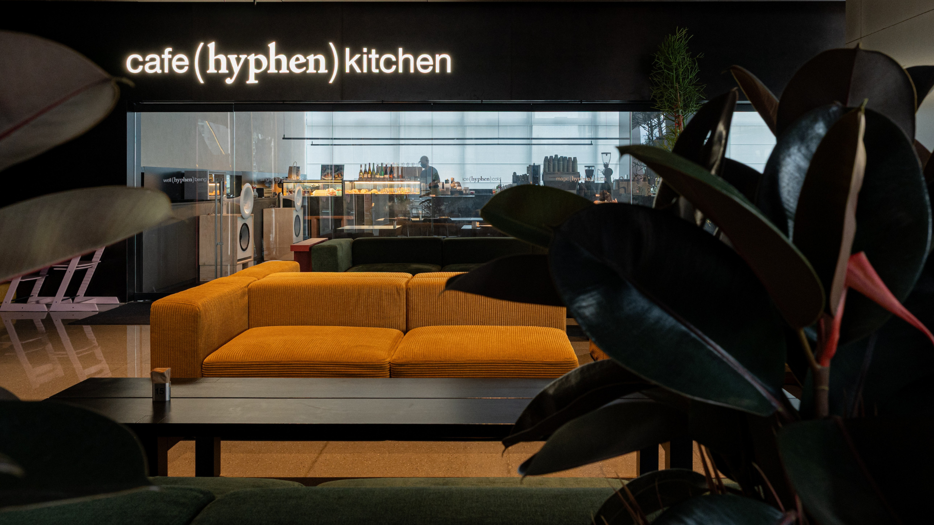 A modern interior scene with a mustard yellow sectional sofa in the center, dark green plants in the foreground, and a black wall sign reading 'cafe(hyphen) kitchen' above a large window overlooking a city skyline at dusk.