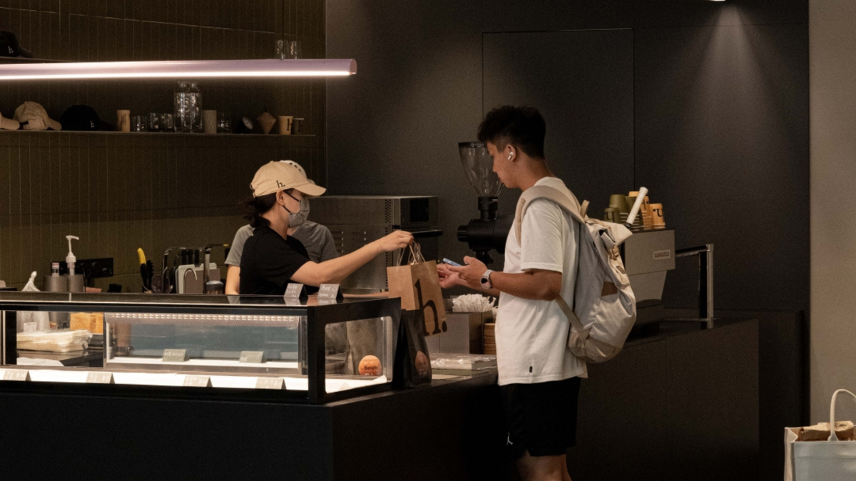 A person in a white t-shirt and black shorts stands at a cafe counter receiving a paper bag from a worker wearing a beige cap and face mask. The background features dark wood paneling, shelves with coffee equipment, and a modern linear light fixture.