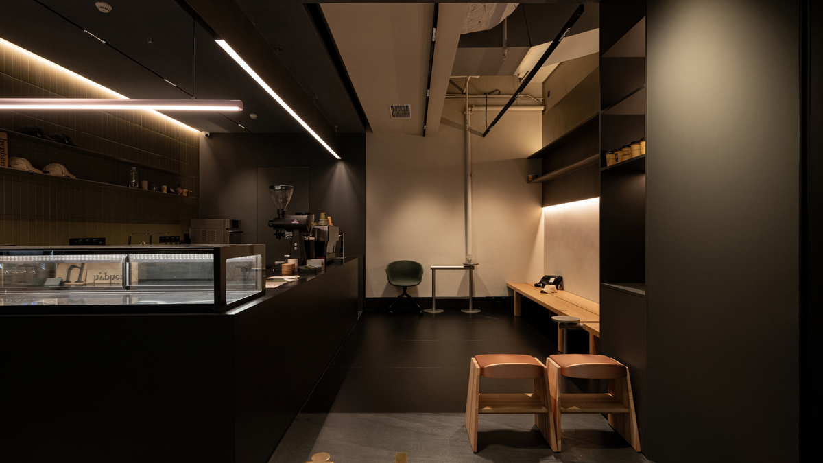 A modern, dimly lit cafe interior with a dark wood counter, a glass display case, a small green chair, wooden stools, and built-in shelving with warm under-shelving lighting.