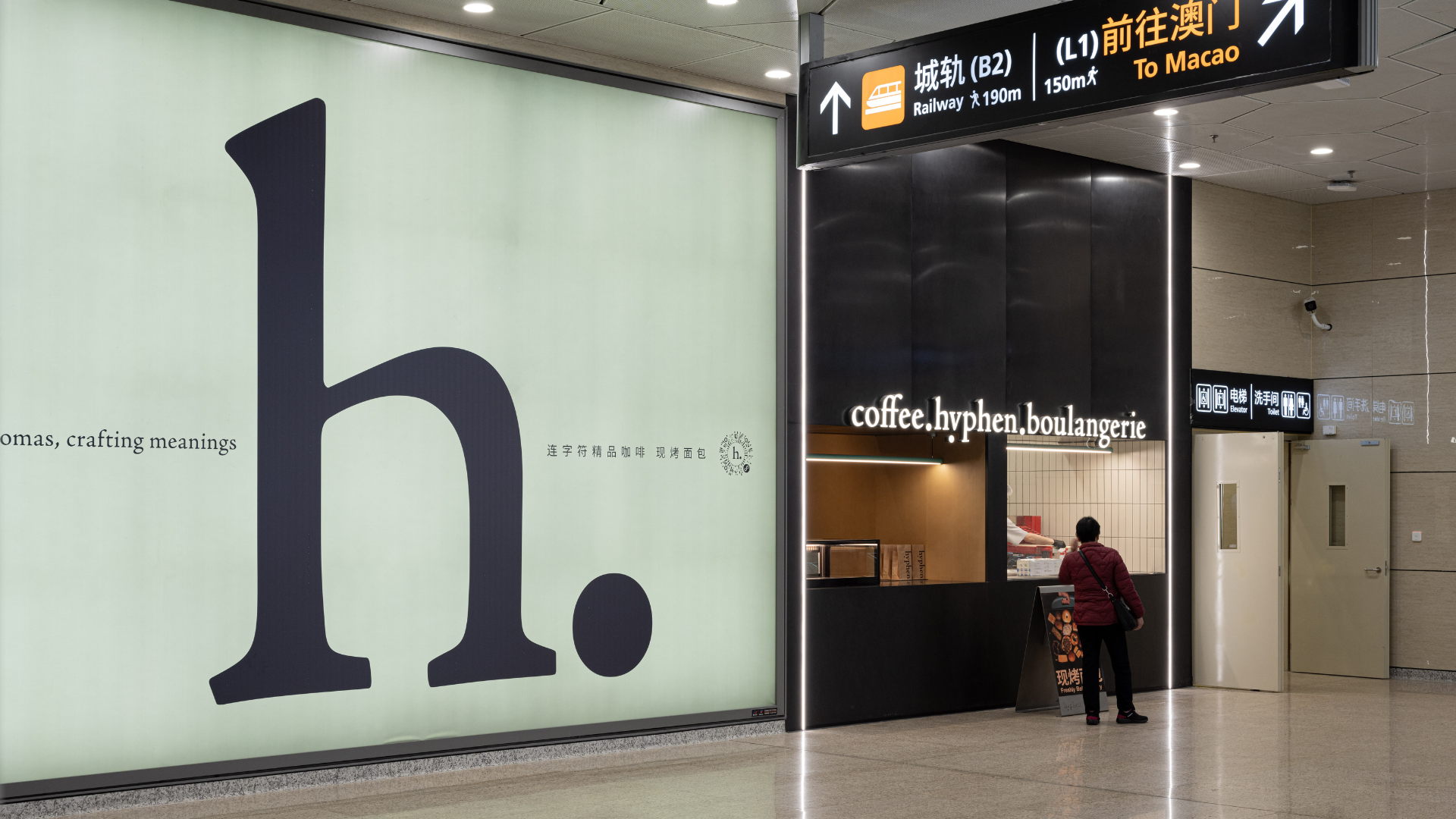 A large minimalist wall sign with a dark navy letter 'h.' on a pale greenish background stands beside a storefront labeled 'coffee.