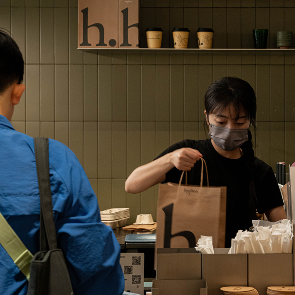 A person in a blue shirt stands at a counter while another person wearing a black shirt and face mask holds a brown paper bag with a large black letter 'h' in a cafe setting with olive-green tiled walls.