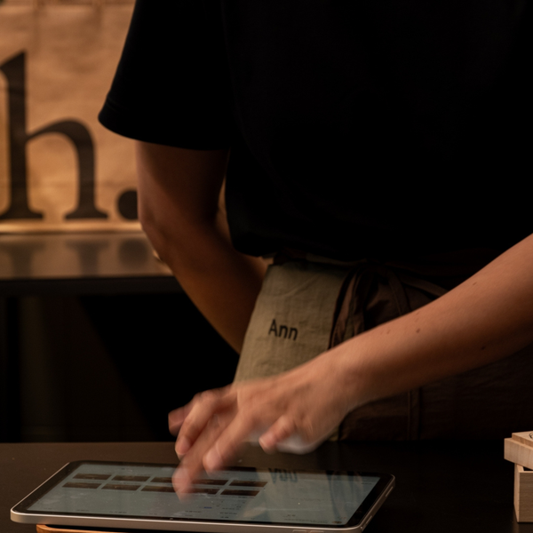A person in a black shirt and beige apron with the name 'Ann' embroidered on it, using a tablet at a counter in a warm, dimly lit restaurant or cafe setting.