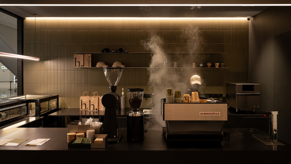 A professional coffee station setup in a dimly lit cafe interior, featuring a La Marzotto espresso machine with steam rising, a coffee grinder, shelves with cups, and warm ambient lighting.