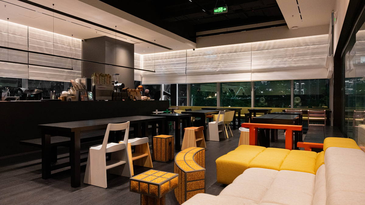 A modern indoor lounge or cafe space with long dark tables, white and yellow seating, wood stools, large windows, and a dark ceiling with linear lighting.