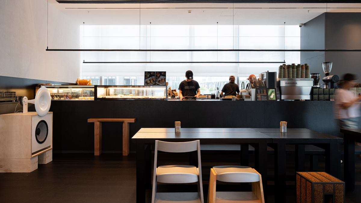 A modern cafe interior with a long black counter, stools, and tables in the foreground, with staff visible behind the counter and large windows in the background.