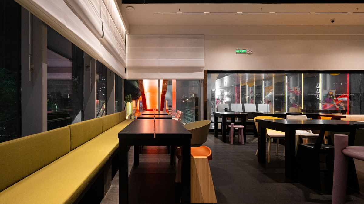 A modern restaurant interior with long dark tables, yellow upholstered booth seating along the left wall, black chairs, large windows, and warm ambient lighting.