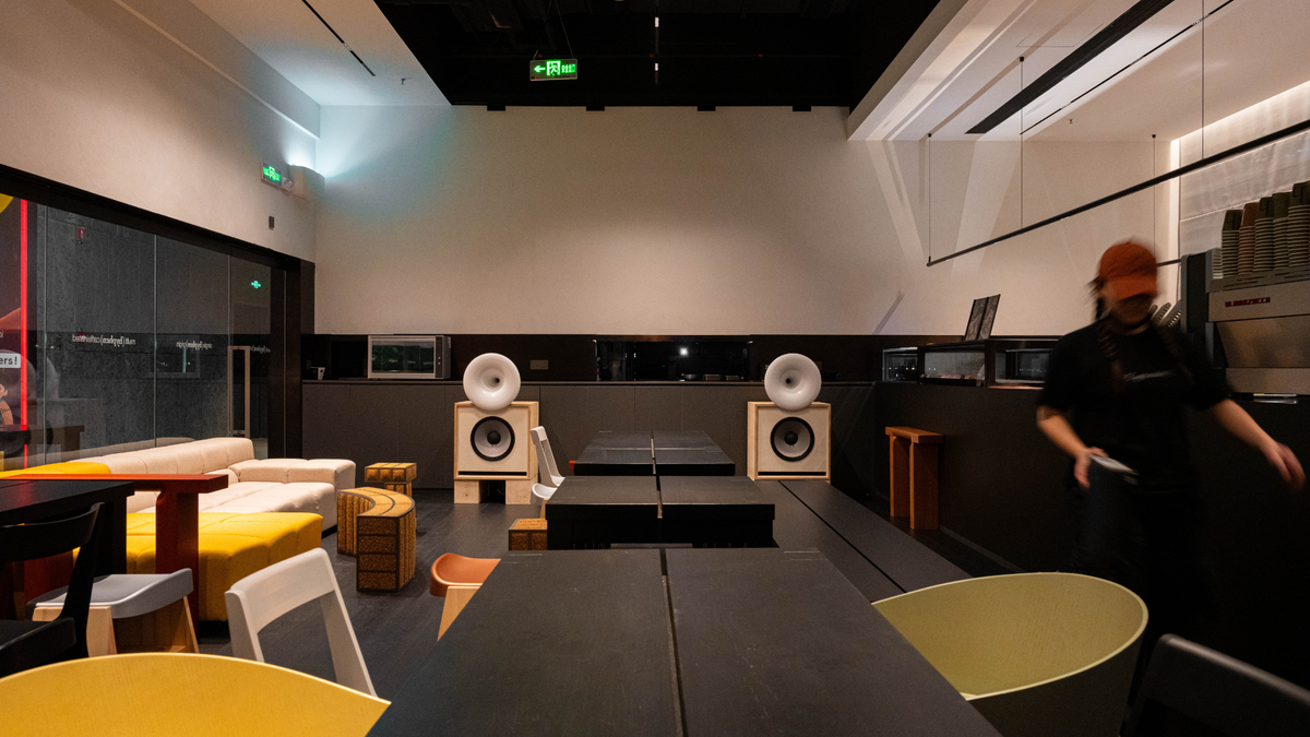 A modern interior space with two large white horn speakers on stands, black tables, yellow and white chairs, shelving, and a person walking in the background.