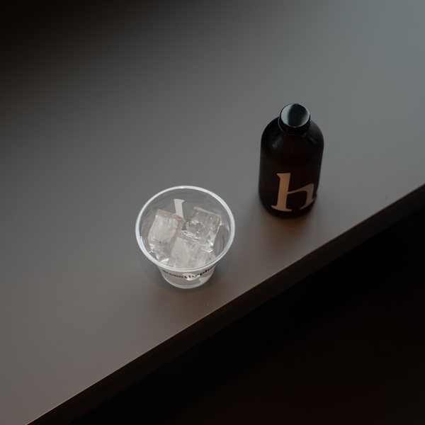 A small clear plastic cup filled with ice cubes sits on a dark brown wooden surface next to a dark brown bottle with a gold letter 'h' on the front.