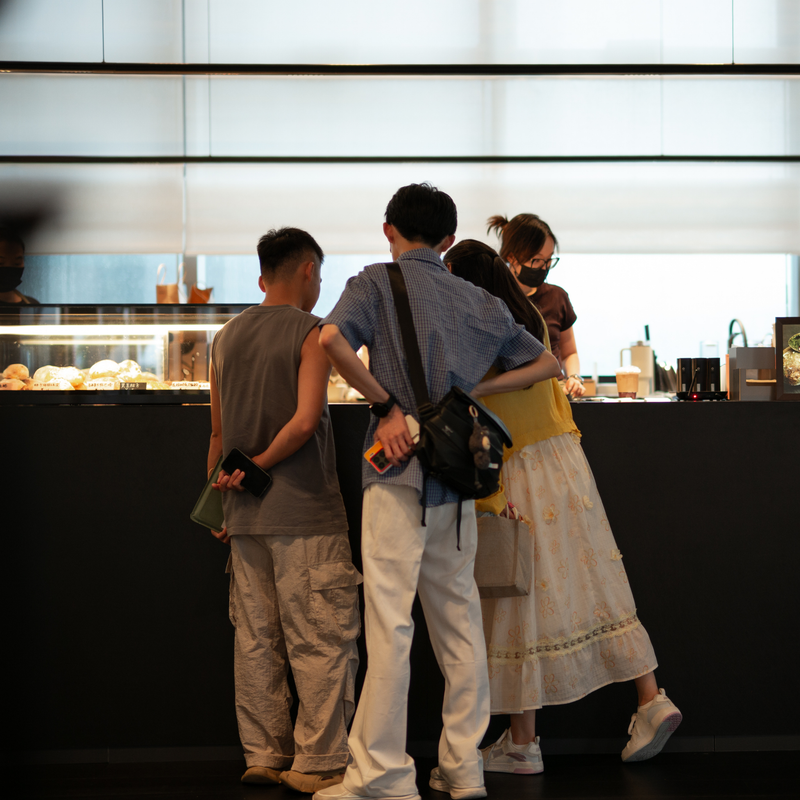 Four people standing in a cafe or bakery, viewed from behind as they face the counter where a staff member is working. The scene is indoors with bright backlighting and a dark counter in the foreground.