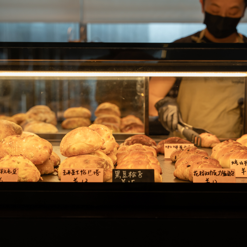 A baker reaching into the lit hyphen. display case, filled with fresh breads and pastries labelled by hand in Chinese.