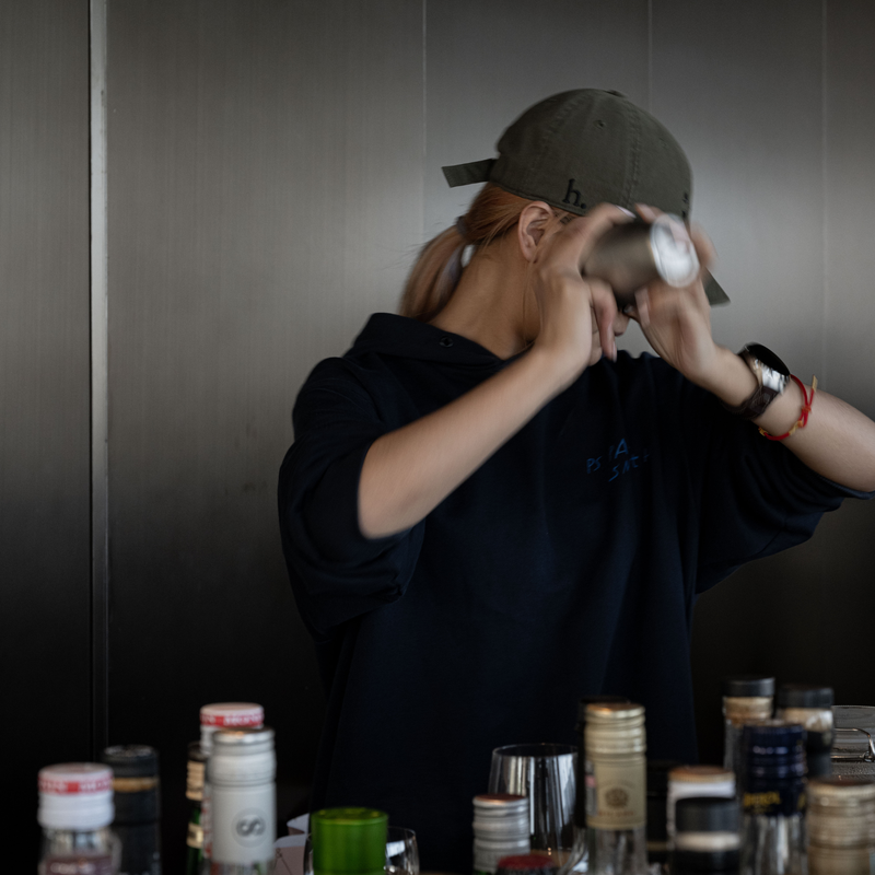 A person wearing a dark navy oversized hoodie and a dark olive green baseball cap is holding a silver metal shaker bottle, captured in motion against a dark gray paneled wall. The foreground shows a table cluttered with various bottles and cups.
