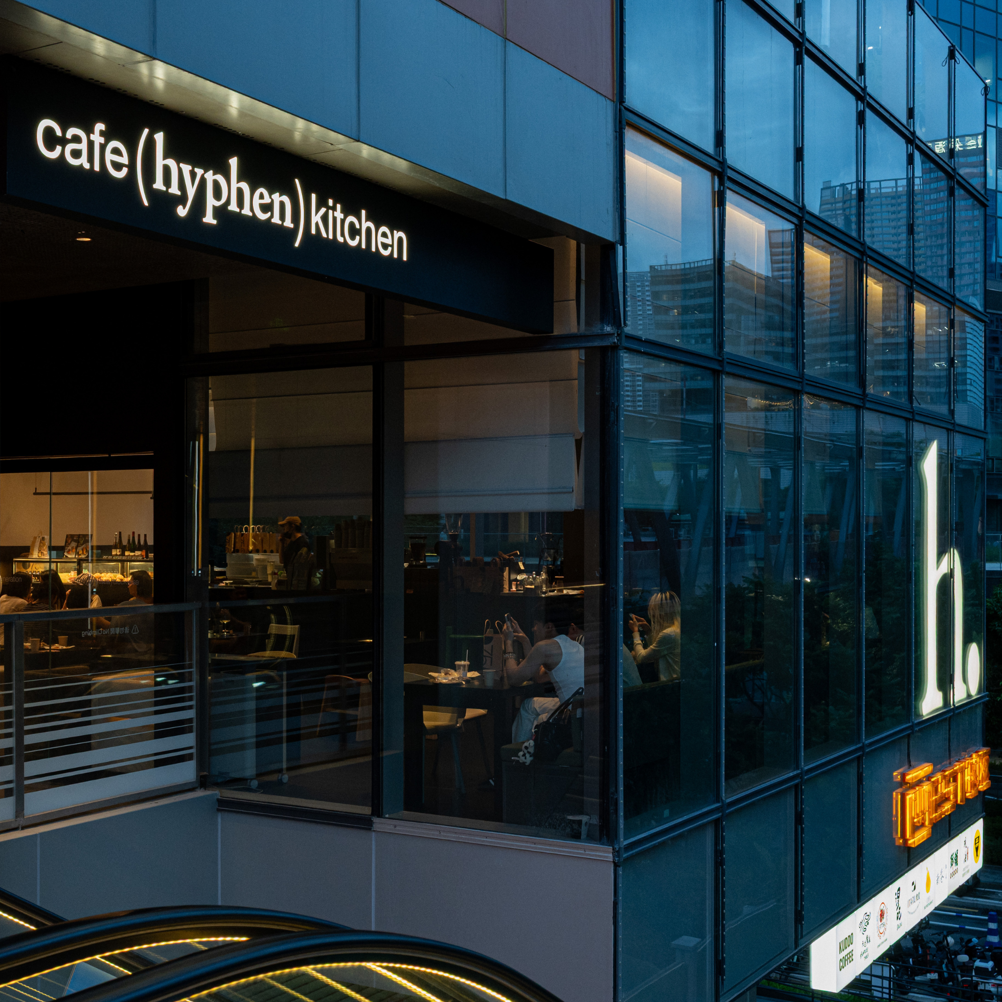 Modern glass-front restaurant exterior with a black sign reading 'cafe (hyphen) kitchen' and a large illuminated white 'h.' on the glass facade at dusk.