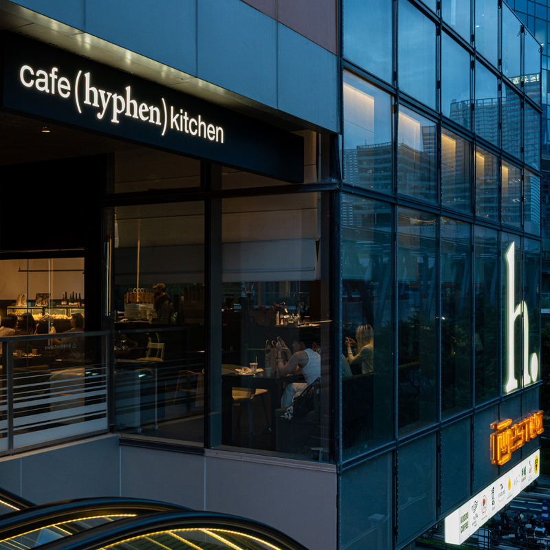 Modern glass-front restaurant exterior with a black sign reading 'cafe (hyphen) kitchen' and a large illuminated white 'h.' on the glass facade at dusk.