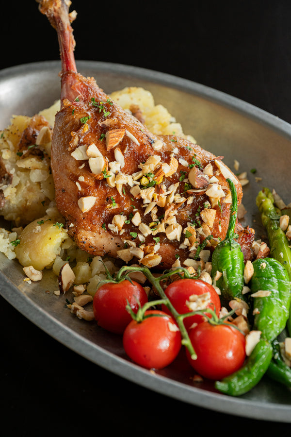 A plated roasted game leg with mashed potatoes, green beans, cherry tomatoes, and chopped nuts on a dark background.