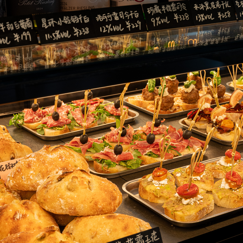 A food display case filled with assorted appetizers and breads, including sliced salami and prosciutto on bread with olives, small meatballs on toothpicks, and bruschetta-style bites with tomatoes and cheese, alongside golden bread loaves in the foreground.