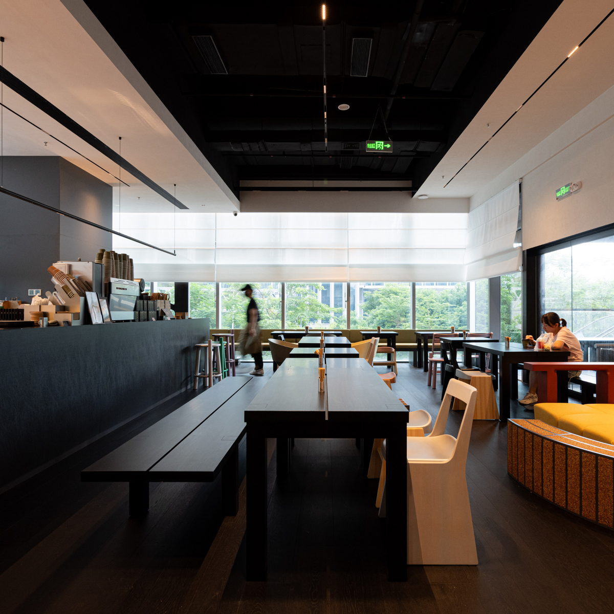 A modern cafe interior with long dark tables and benches, white chairs, a dark service counter, large windows with daylight blinds, and a few seated or walking people in the space.
