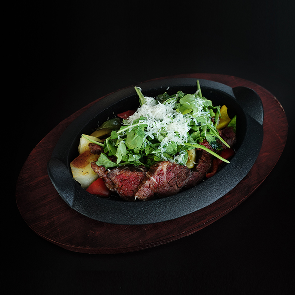 A plated steak dish on a black oval plate with a dark wooden base, topped with greens and shredded cheese, photographed against a black background.