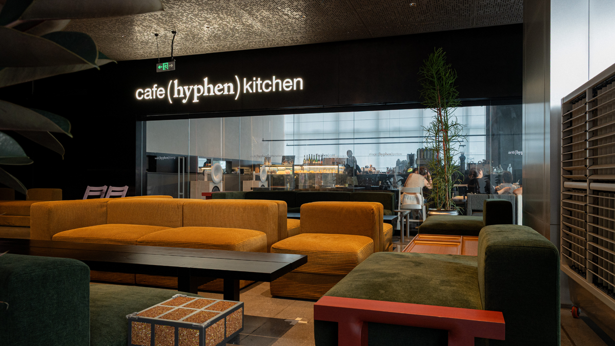 A modern cafe interior with a black wall sign reading 'cafe(hyphen) kitchen' above a glass counter. The seating area features mustard yellow upholstered sofas and green velvet seating with dark wood tables in the foreground.