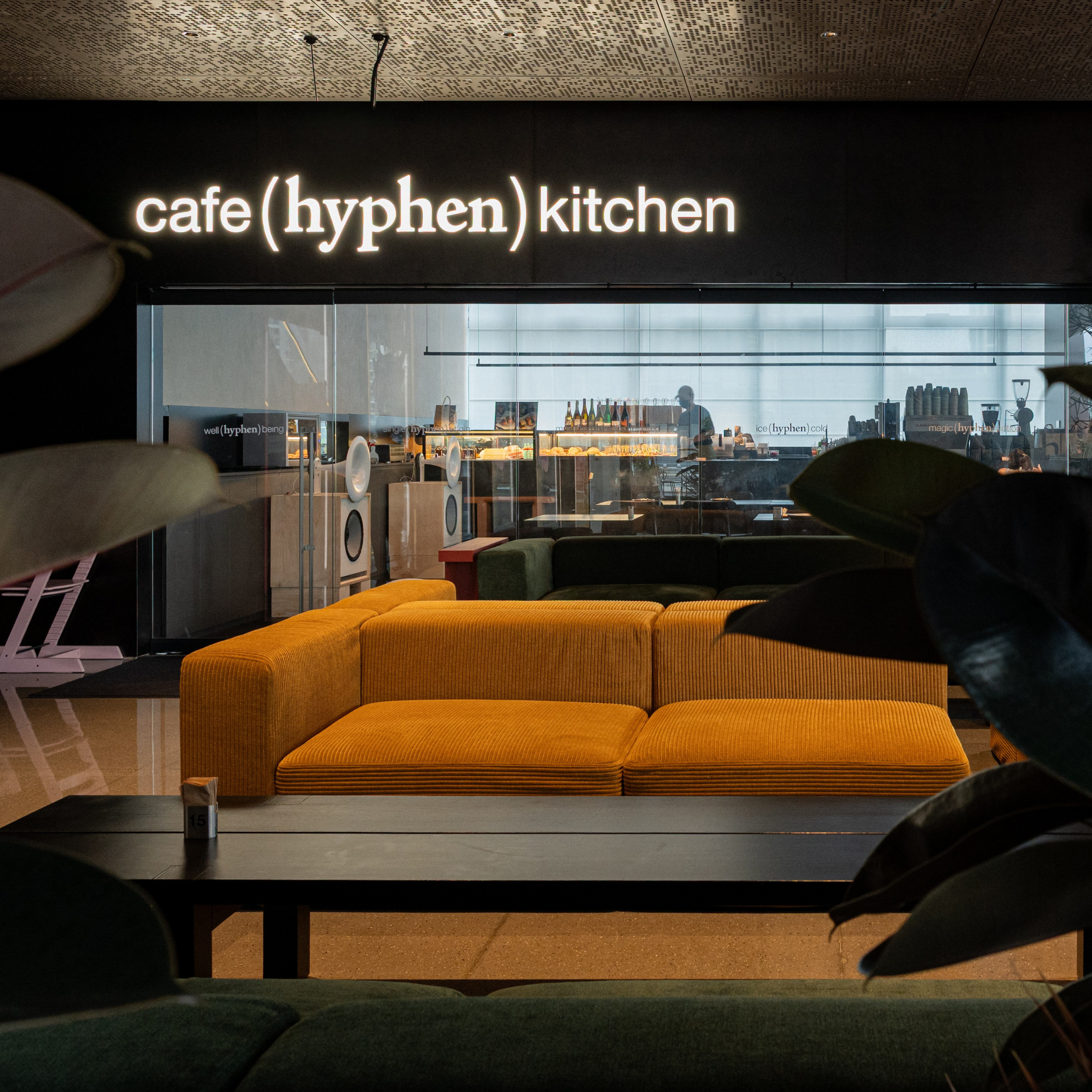 A modern interior scene with a mustard yellow corduroy sofa in the foreground, a dark coffee table, and a large black wall sign reading 'cafe (hyphen) kitchen' above a glass kitchen area. Large green leaves partially frame the foreground.