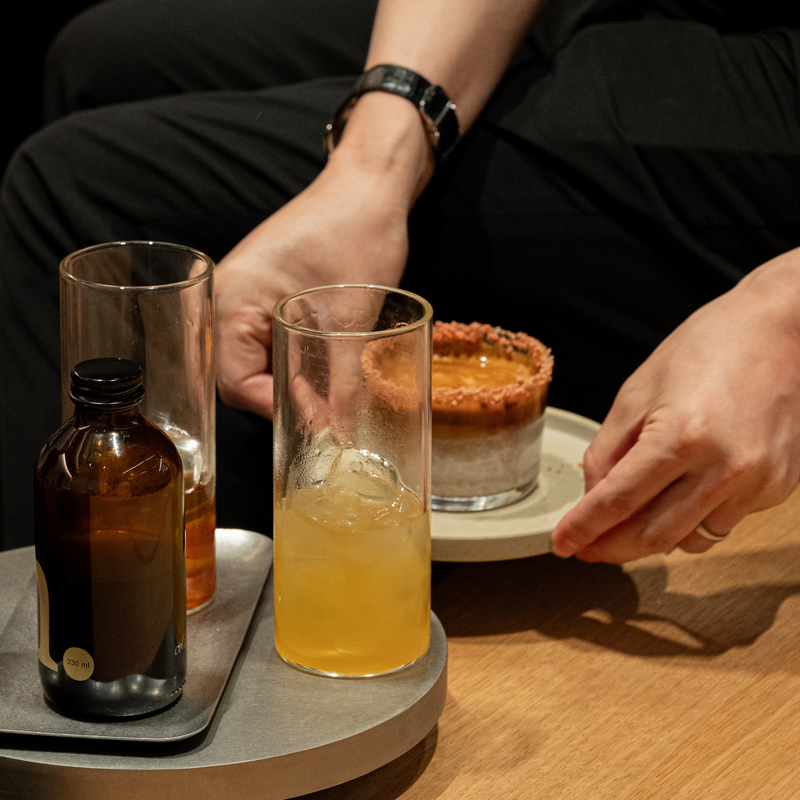 A lifestyle shot of a person's hands and torso as they serve a layered dessert cocktail. In the foreground, a clear glass filled with an orange-yellow drink sits on a gray tray next to an amber glass bottle.