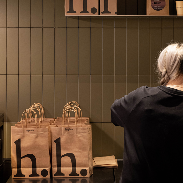 A person with light hair seen from behind stands at a counter next to several brown paper shopping bags printed with the h.h.