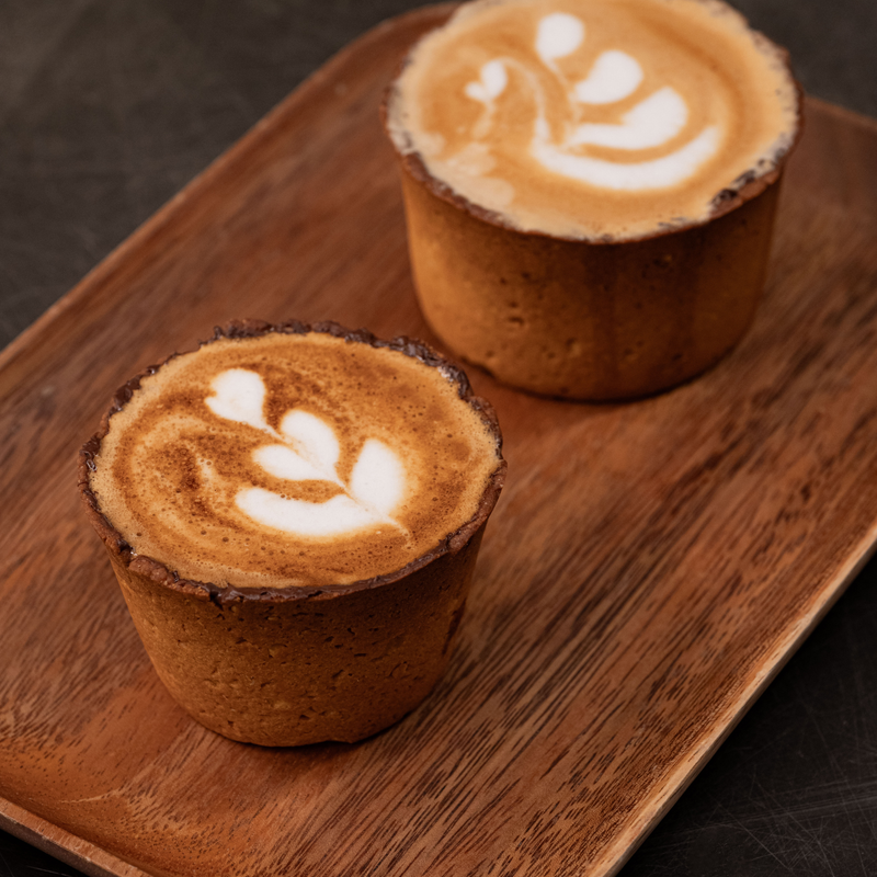 Two small cups of coffee with latte art, served on a rectangular wooden tray against a dark background.