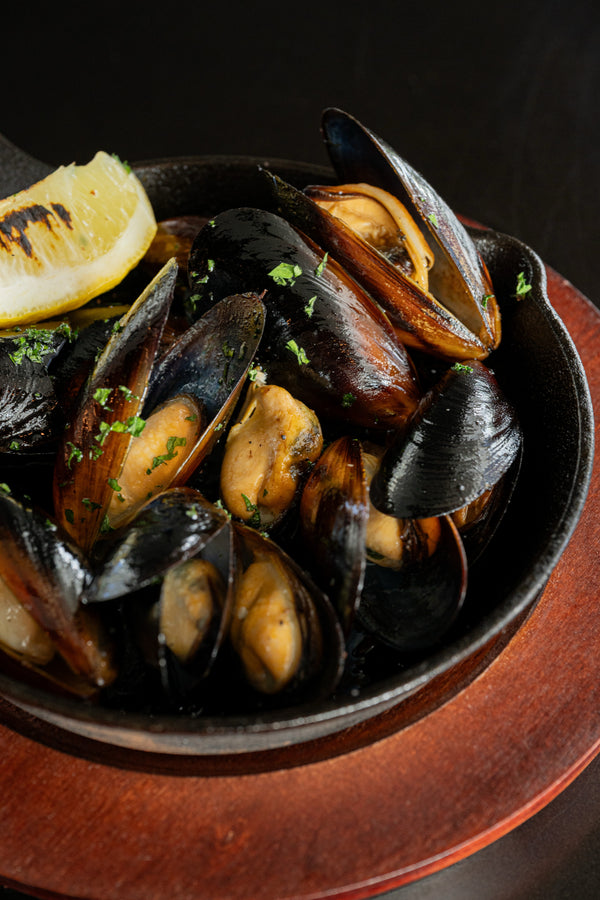 Mussels cooked in a dark cast-iron skillet with a lemon wedge and chopped green herbs, presented on a wooden plate against a dark background.