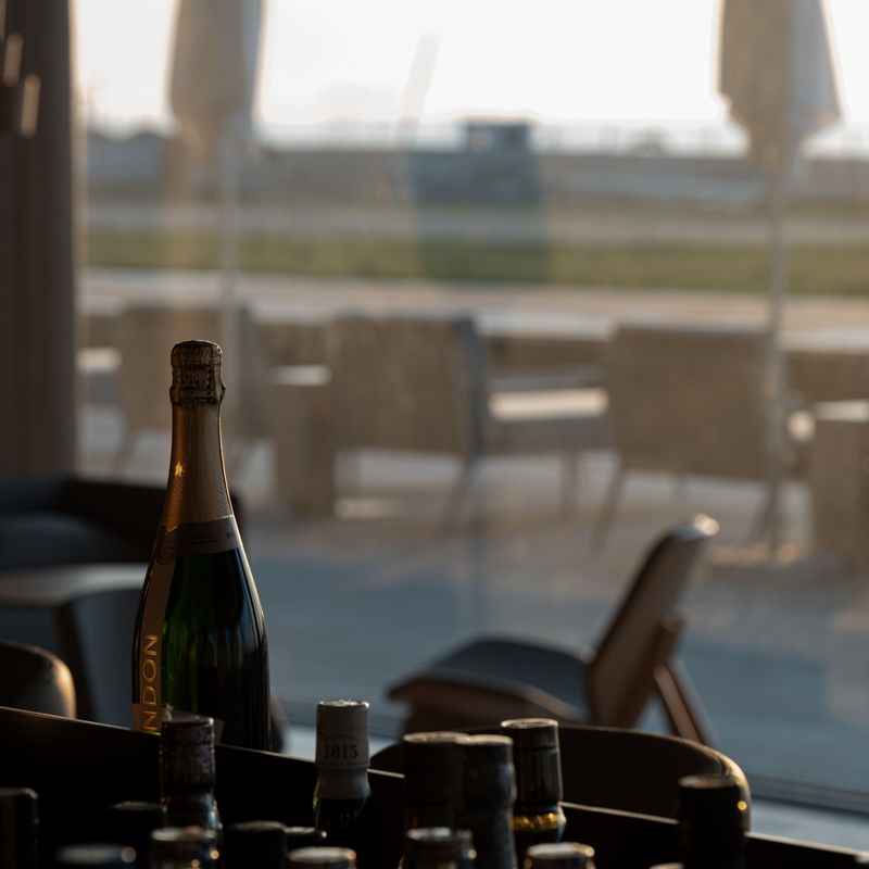 A dark green bottle of Noni sparkling wine or champagne with a gold foil label, standing on a dark table in a dimly lit setting with a blurred outdoor patio and lounge chairs in the background.